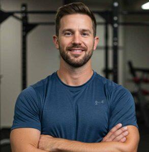 A friendly, confident American male fitness coach in his late 30s, light skin tone, short brown hair, light stubble beard, wearing a fitted athletic shirt (dark blue or black), standing in a gym environment with blurred barbells and squat rack in the background. Natural lighting, slight smile, arms crossed, clean and professional look. No logos or brands on clothing.